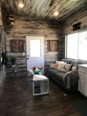 A rustic interior with a wooden ceiling, a white door, a grey sofa with patterned cushions, a small table with a bowl, and a window with a view.