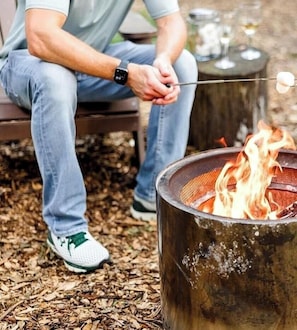 A person sitting by a metal fire pit with a stick holding a marshmallow. - Stunning Tented Cabin with Fire Pit in Des Moines, Iowa (Des Moines)