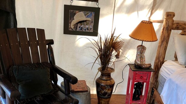A rustic room with a wooden rocking chair, a small red table with a lamp, a vase with dried plants, and a framed picture on the wall. - Pet-Friendly Safari Tent Glamping in Swan Valley, Idaho near Jackson Hole (Irwin)