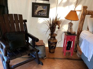 A rustic room with a wooden rocking chair, a small red table with a lamp, a vase with dried plants, and a framed picture on the wall.