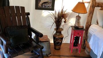 Une pièce rustique avec un fauteuil à bascule en bois, une petite table rouge surmontée d’une lampe, un vase contenant des plantes séchées et un tableau encadré accroché au mur.