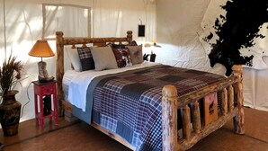 A rustic wooden bed with a checkered bedspread, a small red nightstand with a lamp, and a cowhide wall. - Pet-Friendly Safari Tent Glamping in Swan Valley, Idaho near Jackson Hole (Irwin)