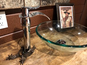 Une salle de bain avec un lavabo en verre, un robinet et un tableau encadré au mur.