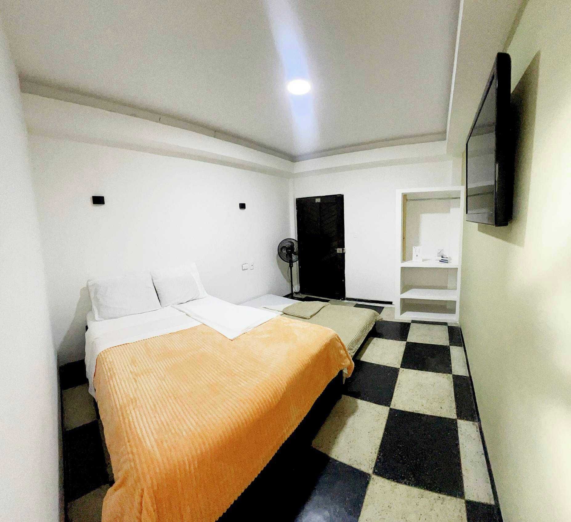 A hotel room with a bed, a fan, a wardrobe, and a TV mounted on the wall.