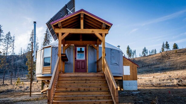 Une cabane en bois avec une porte rouge, un escalier menant à l’entrée et un panneau solaire sur le toit.