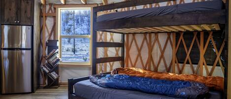 Interior acogedor de una cabaña con literas, un refrigerador, una estantería y una ventana con vistas a la nieve.