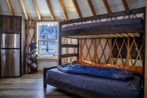 Un intérieur de chalet chaleureux avec un lit superposé, un réfrigérateur, une bibliothèque et une fenêtre donnant sur un paysage enneigé.