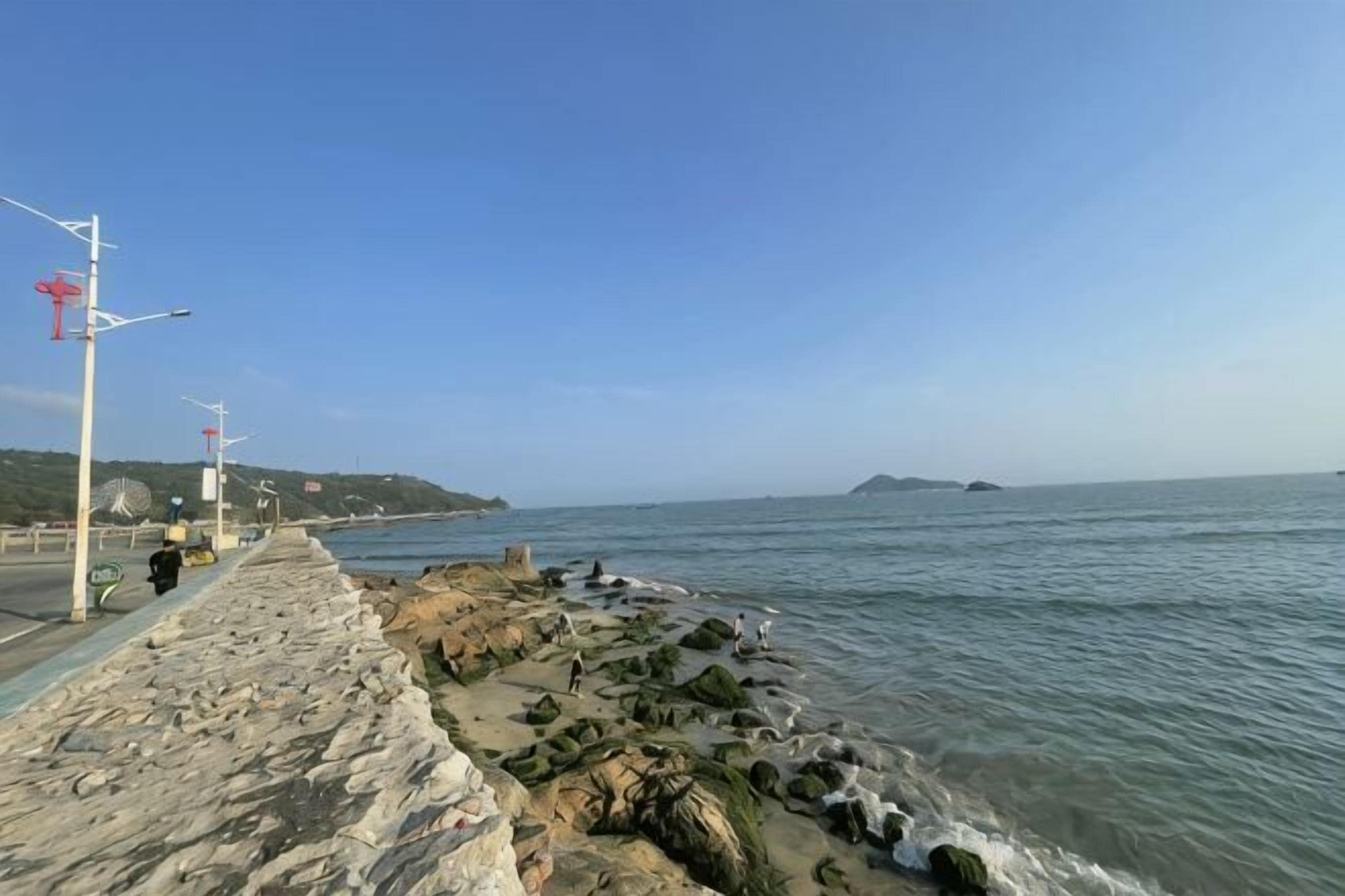 Un paseo marítimo con muro de piedra, cielo despejado y mar en calma.