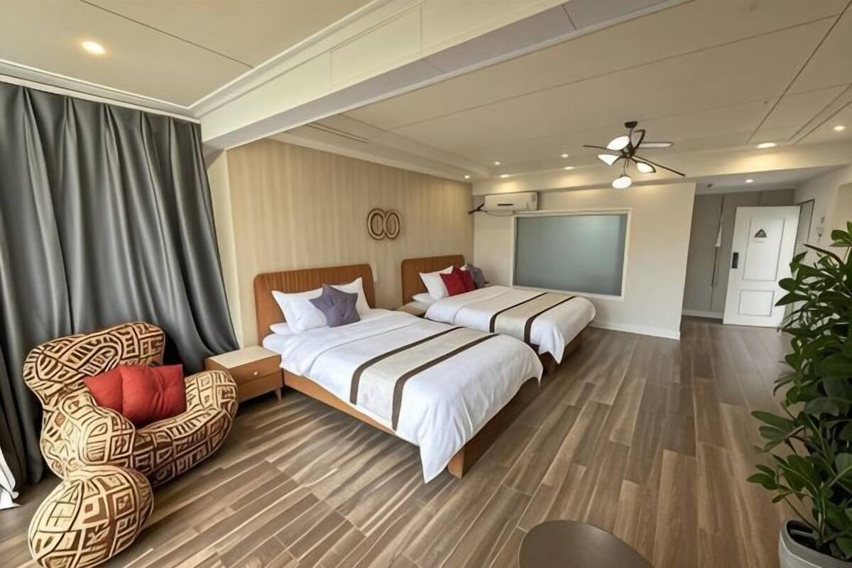 Hotellihuone, jossa on kaksi sänkyä, kuviollinen nojatuoli, viherkasvi ja moderni kattotuuletin.