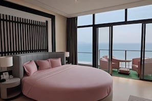 Een groot bed met hoofdeinde, nachtkastjes met lampen en uitzicht op zee door grote ramen.
