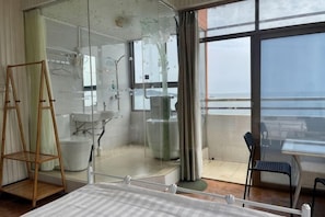 A modern bathroom with a glass shower enclosure, a sink, and a toilet. There is a bed with a white bedspread, a chair, and a table with a view of the sea.