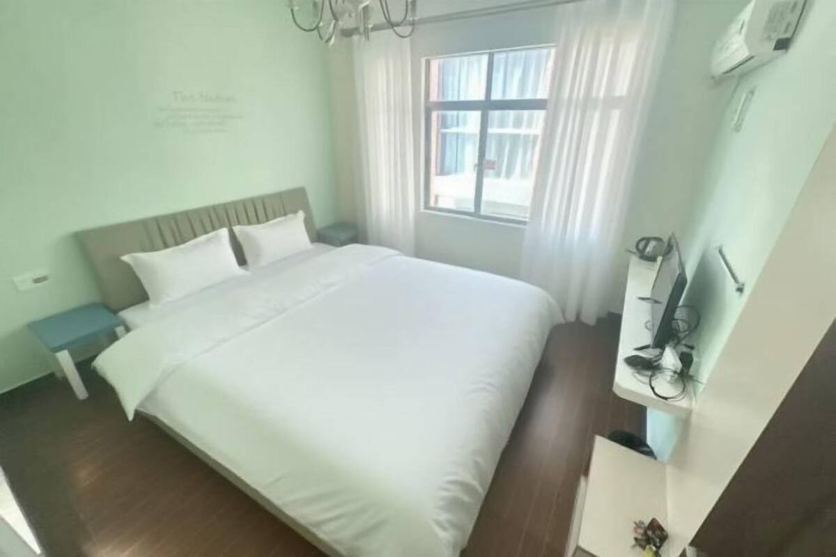 Hotellihuone, jossa on sänky, yöpöytä, ikkuna verhoilla ja seinälle asennettu ilmastointilaite.