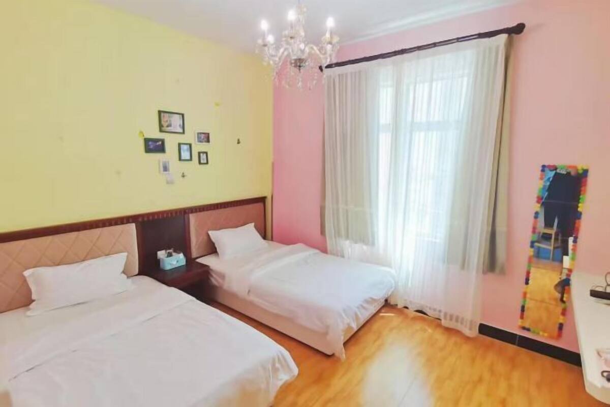 Hotellihuone, jossa on kaksi sänkyä, kattokruunu, ikkuna läpikuultavilla verhoilla ja seinä, jossa on kehystettyjä tauluja.
