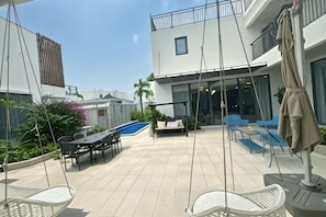 Een modern terras buiten met een eettafel, stoelen, een hangstoel, een zwembad en lounge meubels.