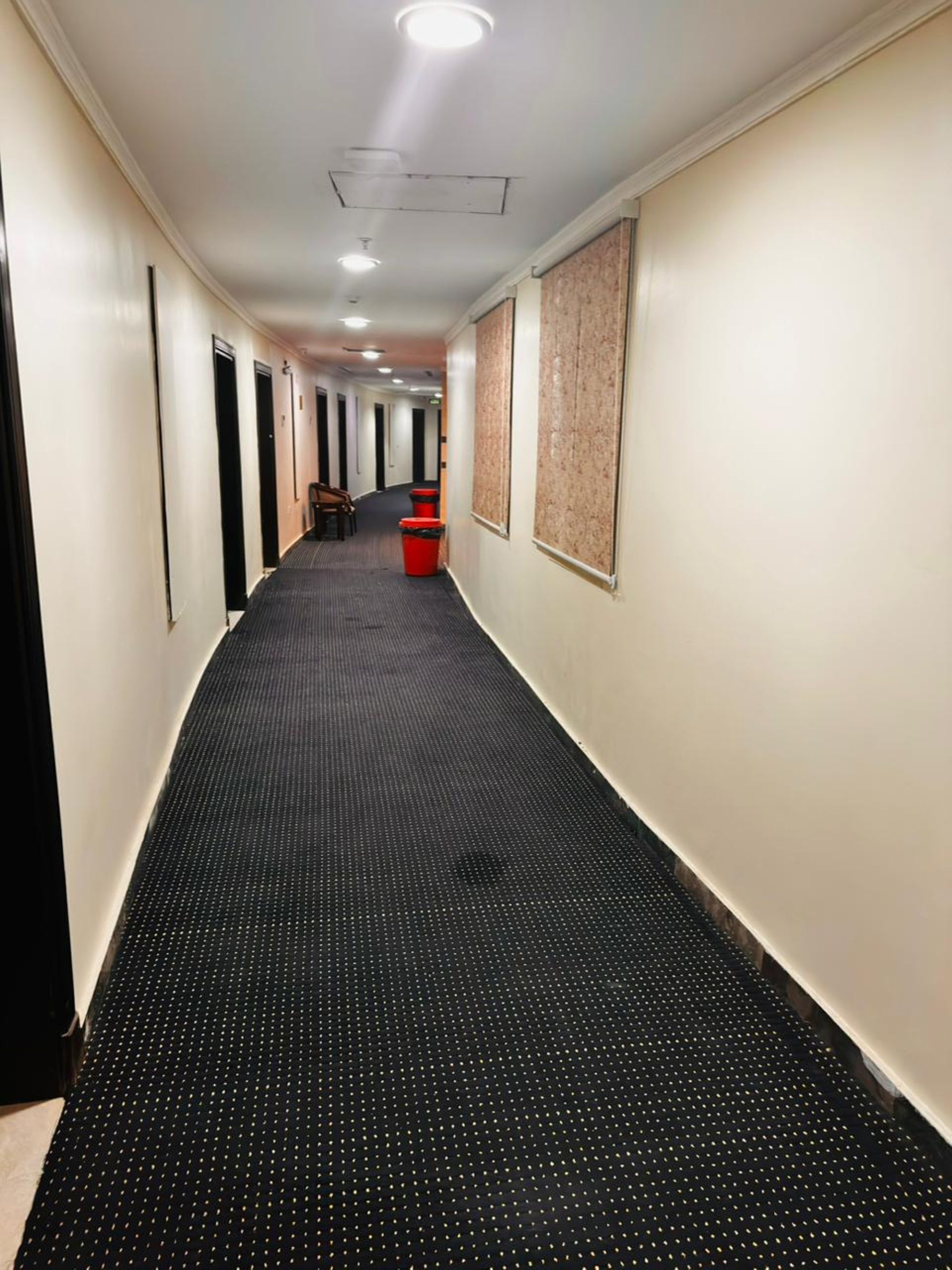 Un long couloir étroit, doté d’un tapis noir à motifs pointillés, de murs blancs et de portes de part et d’autre.