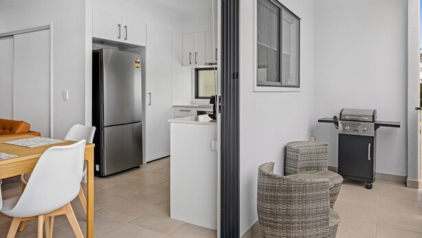 A compact kitchen with a refrigerator, microwave, and grill, a dining area with chairs and a table, and a window with blinds. - Riverside 3-bed Home With Study Nook and BBQ (Murrumba Downs)