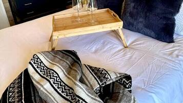 A bedroom with a bed, a wooden bedside table with glasses, a mirror, a window, and a wall-mounted air conditioner.