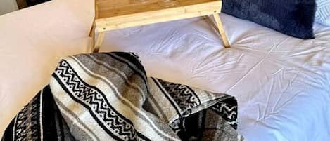 A bedroom with a bed, a wooden bedside table with glasses, a mirror, a window, and a wall-mounted air conditioner.