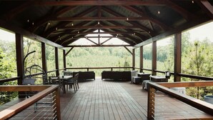 Una terraza techada al aire libre con piso de madera, arreglos de asientos y ventanas amplias que ofrecen vistas de la vegetación circundante.