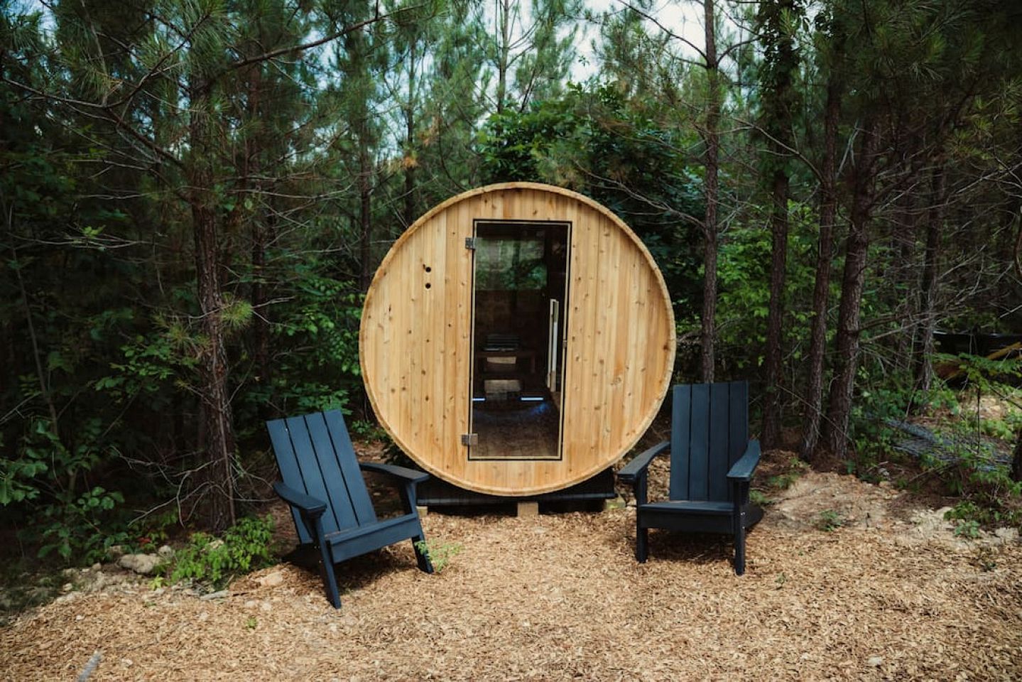 A wooden sauna in a forest with two Adirondack chairs.