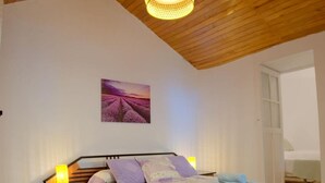Une chambre avec un plafond en bois, un lit recouvert d’une courtepointe blanche et violette, une table de chevet avec une lampe, et un tableau représentant des champs de lavande accroché au mur.