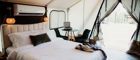 Una cama con cabecera, un equipo de aire acondicionado, una mesita pequeña y una silla en una habitación con forma de tienda.