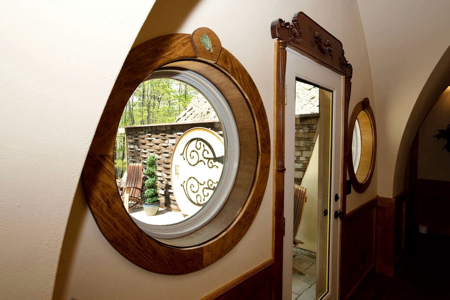 A room with a circular window, a mirror, and a door leading to an outdoor area with a brick wall and a wooden chair.