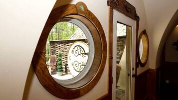A room with a circular window, a mirror, and a door leading to an outdoor area with a brick wall and a wooden chair.
