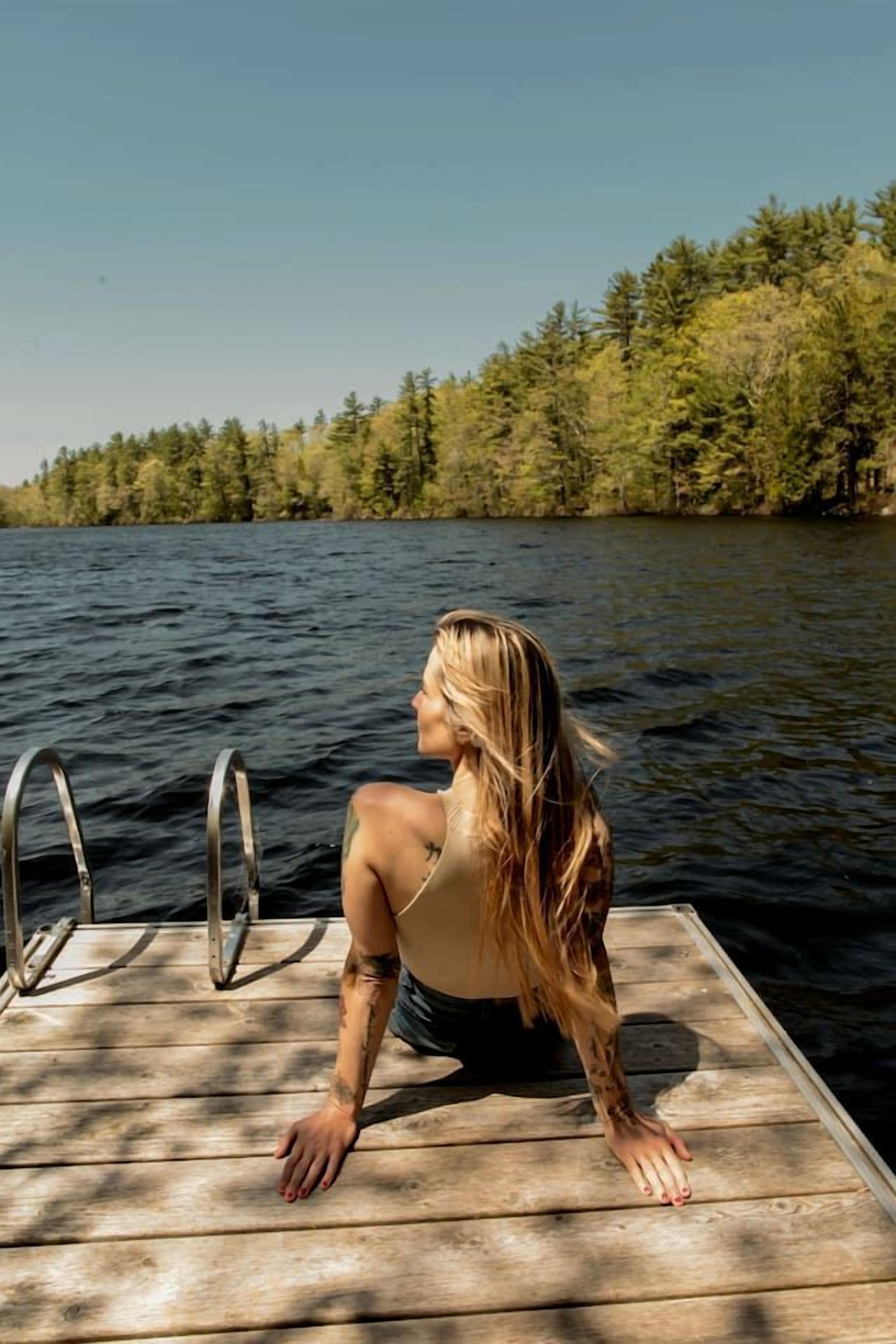 Una persona sentada en un muelle de madera que se extiende sobre el agua, con árboles al fondo.