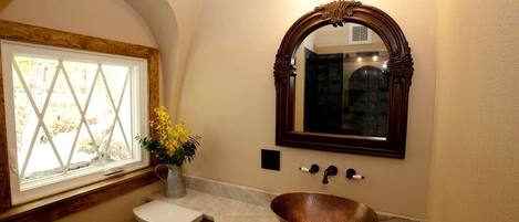 Un baño con inodoro blanco, lavabo de estilo cobre, un florero con flores amarillas, ventana con marco de madera y espejo con arco decorativo.