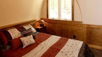 A bedroom with a sloped ceiling, a bed with multiple pillows, a bedside table with a lamp, and a window with a view of trees.