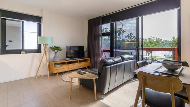 A modern living room with a leather sofa, a wooden coffee table, a TV on a wooden stand, and large windows offering a view of the outdoors. - Comfy 2-bedroom Apartment With a Clyde River View (Batemans Bay)