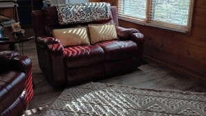A cozy living room with a leather sofa, a patterned rug, and a large window with blinds. - Honey Bear Hideway - Hot Tub - Close to Blue Ridge and Ellijay (Ellijay)