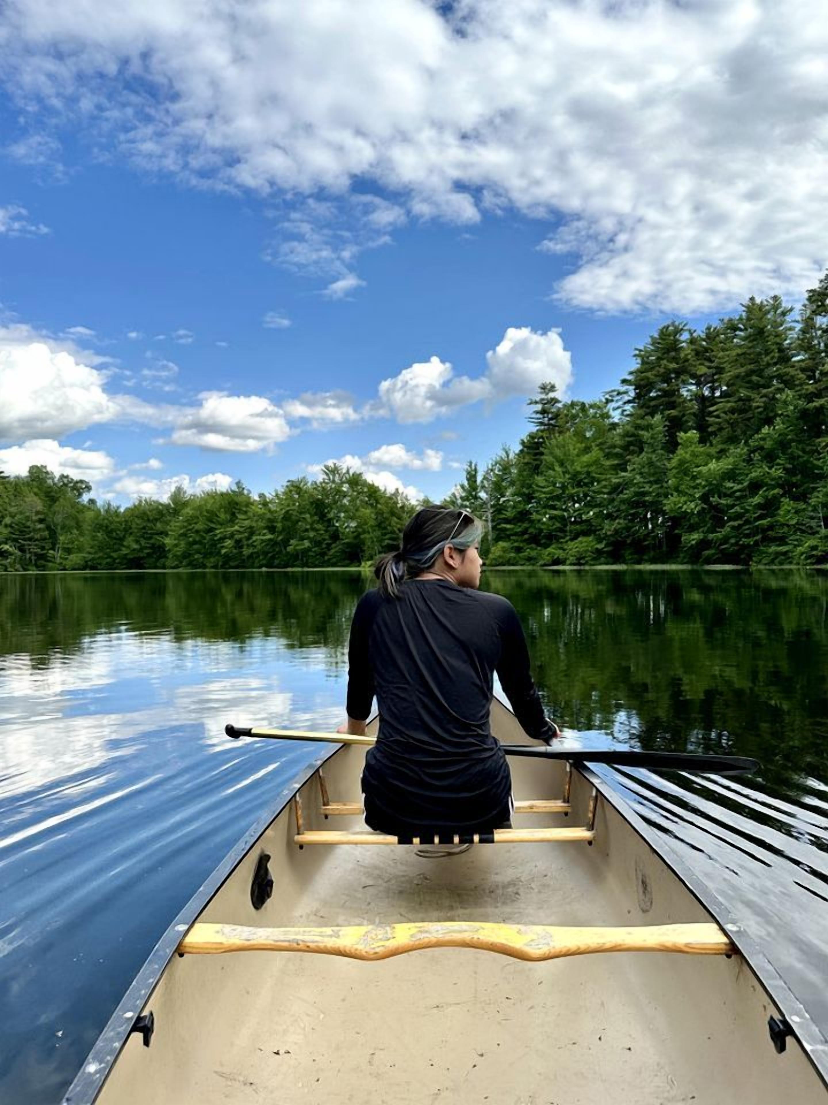 Eine Person sitzt in einem Kanu auf einem ruhigen See, im Hintergrund sind Bäume zu sehen.