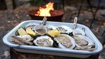 A wooden barrel with a white tray containing oysters, lemon wedges, and a small bowl of sauce, set outdoors with a fire in the background.