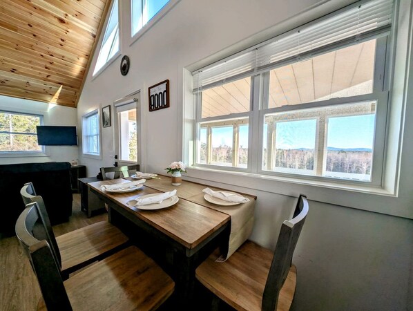 A dining area with a wooden table, chairs, and a view of the outdoors through large windows.