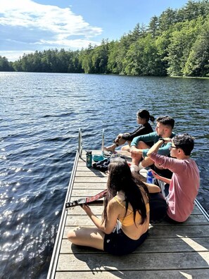 Un grupo de personas sentadas en un muelle de madera a orillas de un lago, con árboles al fondo.