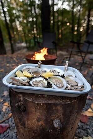 Un tonneau en bois surmonté d’un plateau blanc contenant des huîtres, des quartiers de citron et un petit bol de sauce, le tout installé à l’extérieur, avec un feu de camp en arrière-plan.