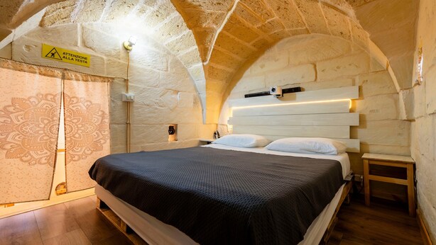 Een slaapkamer met een stenen plafond, een bed met een zwart dekbed, een houten nachtkastje en een wandlamp.