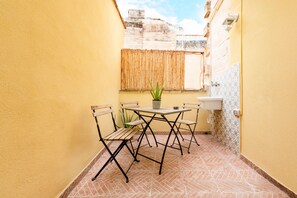 A small outdoor patio with a table and chairs, a potted plant, and a window with a bamboo blind.