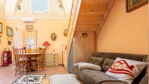 Een knus vertrek met een houten plafond, een eettafel met stoelen, een bank met kussen, en een wand met ingelijste foto's.