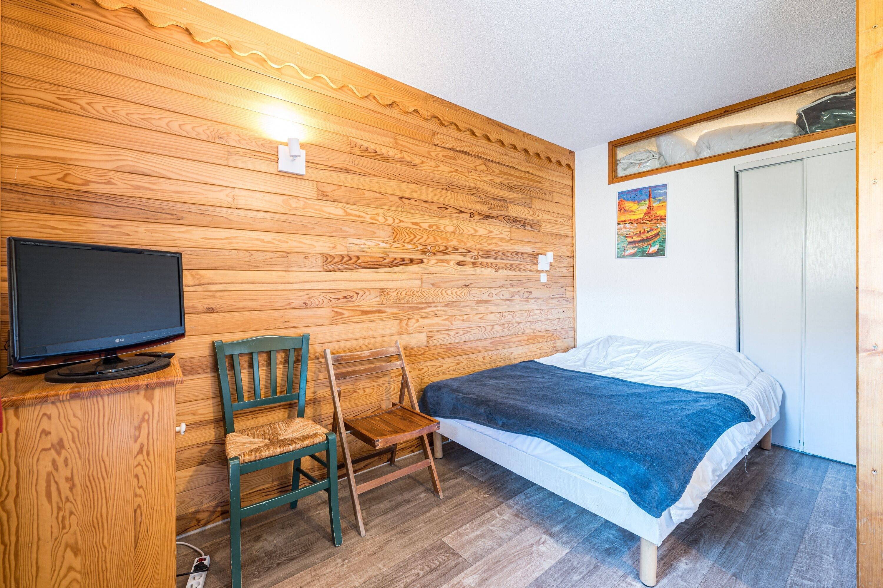 A bedroom with a wooden wall, a bed, a chair, a TV, and a painting.