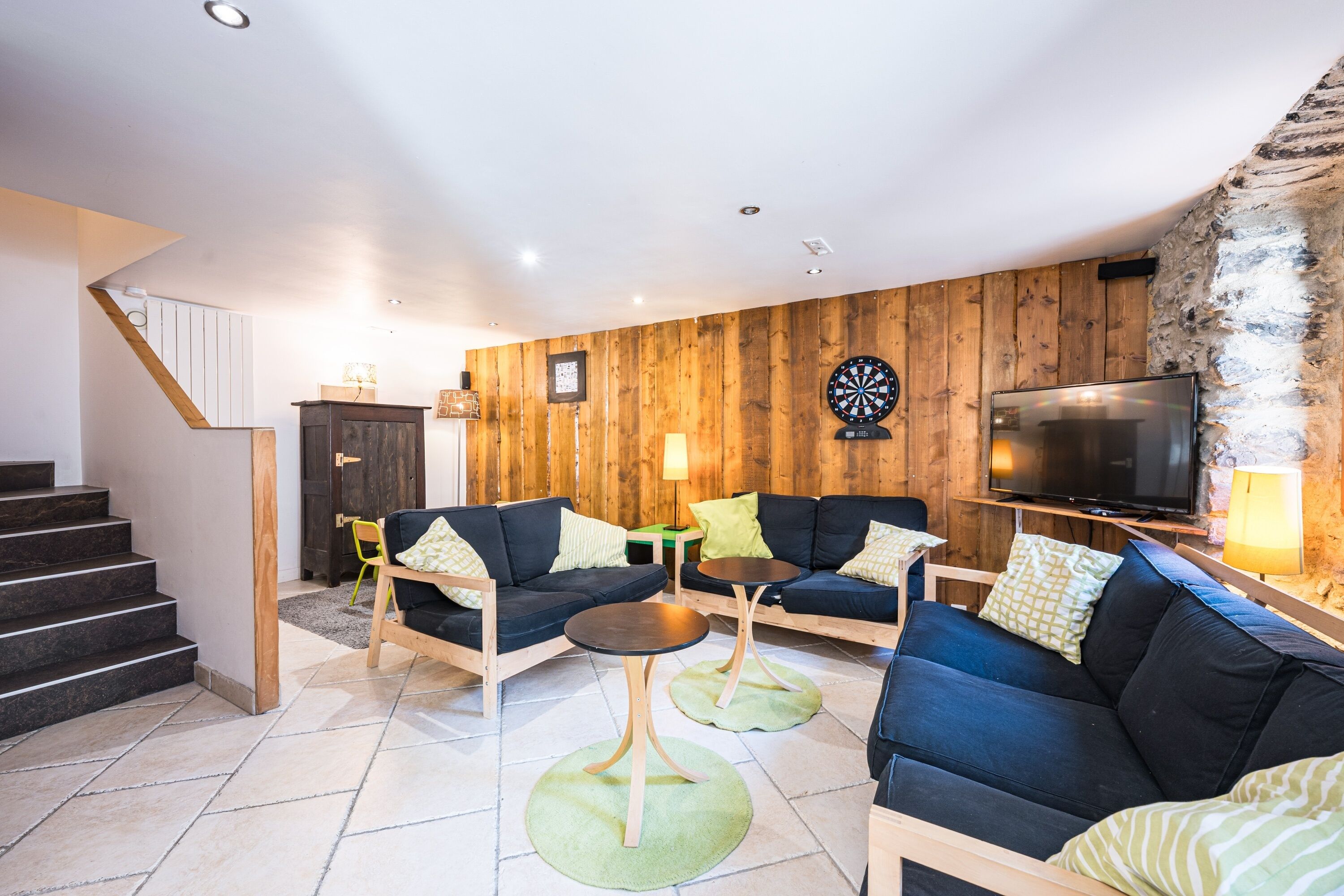 Een moderne woonkamer met een trap, houten wanden, een flatscreen televisie en twee banken met groene kussens.