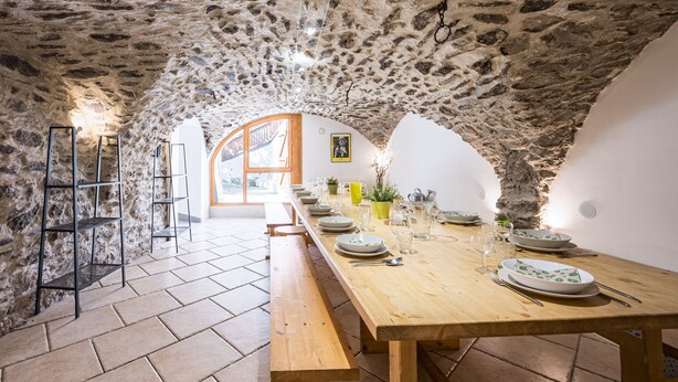 A stone-walled dining area with a long wooden table set for a meal, benches along the walls, and a view through an arched doorway. - La Maison - Chalet DDA La Combe (Saint Martin de Belleville)