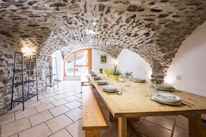 A stone-walled dining area with a long wooden table set for a meal, benches along the walls, and a view through an arched doorway. - La Maison - Chalet DDA La Combe (Saint Martin de Belleville)