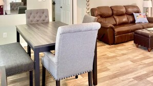 Ein Essbereich mit einem Holztisch, grau gepolsterten Stühlen, einem braunen Ledersofa und einem Holzboden mit Fischgrätmuster.