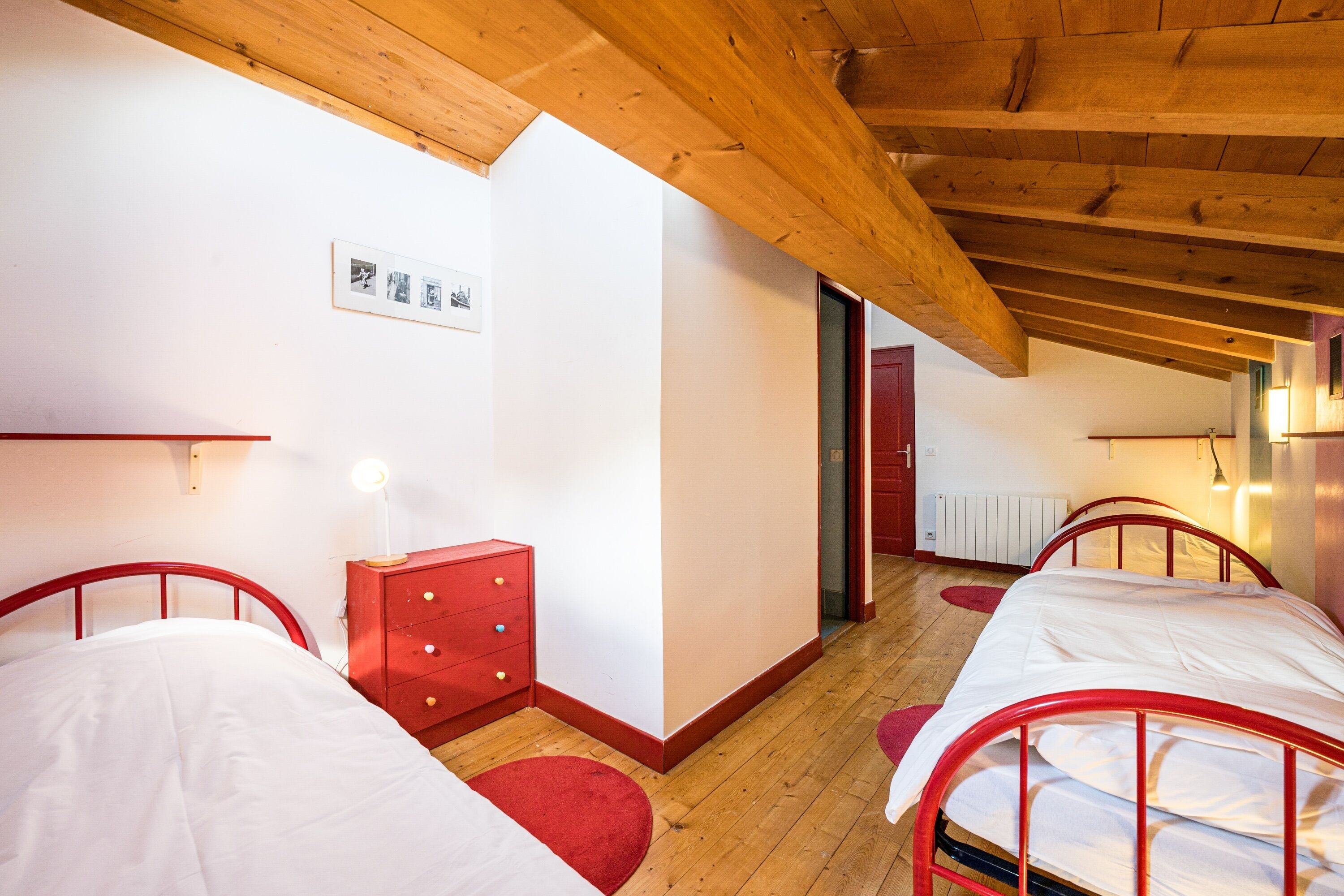 A room with two beds, a red dresser, a wooden ceiling, and a white wall with a framed picture.