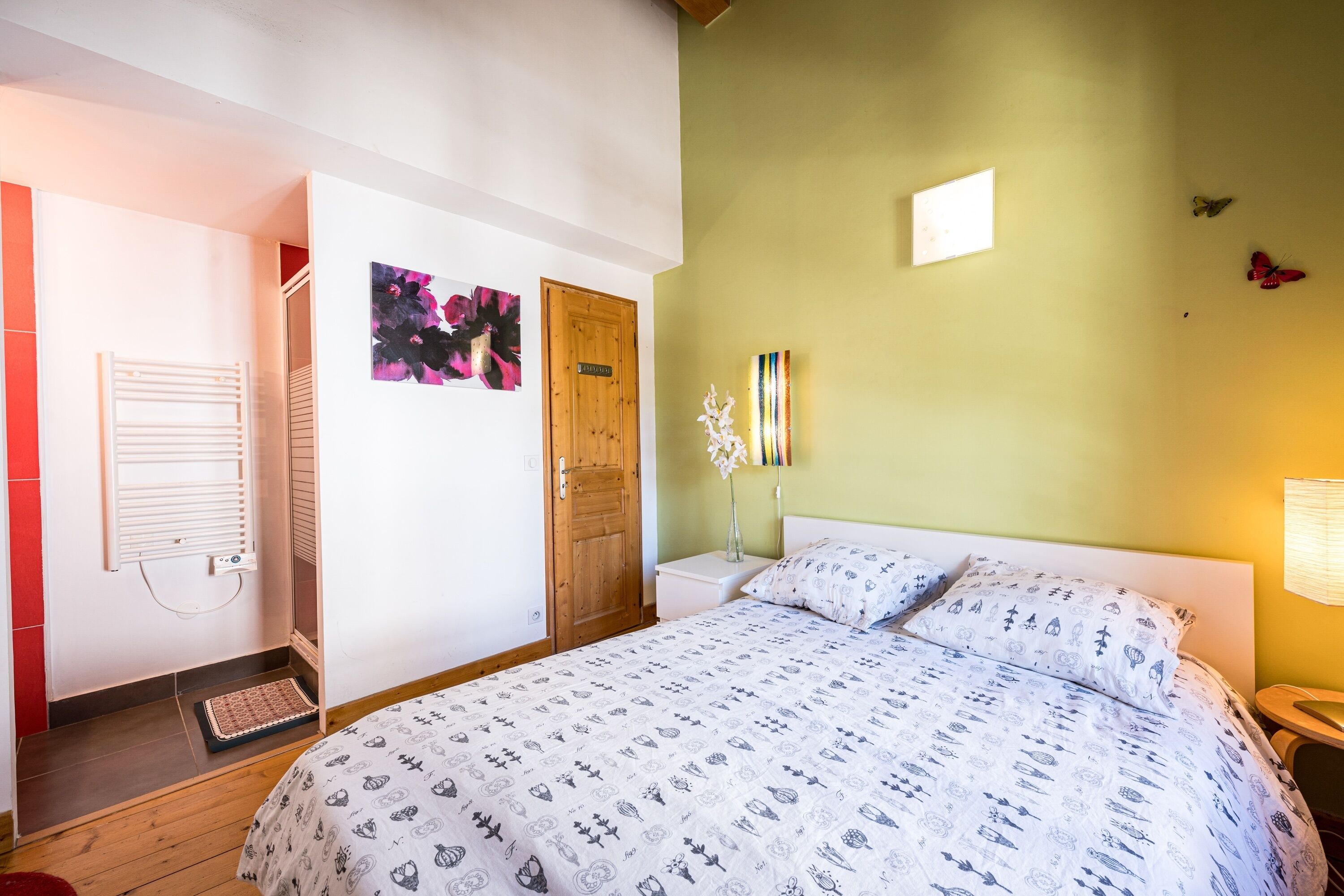 A bedroom with a bed, a wooden door, a wall-mounted light, and a small table with a vase.