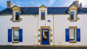 Un edificio blanco con puerta y contraventanas azules, tres ventanas y un pequeño balcón.