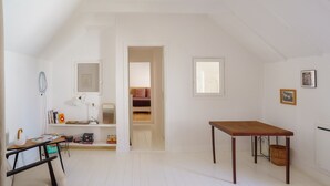 Una habitación amplia con techo alto, una mesa de madera, un escritorio con silla, una estantería y un vano que da a otra habitación.